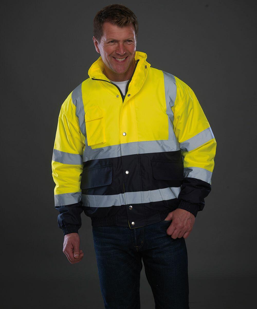 A man is wearing a high-visibility yellow and black Yoko YK044 jacket with reflective stripes. He has a slight smile and is standing against a plain dark background.