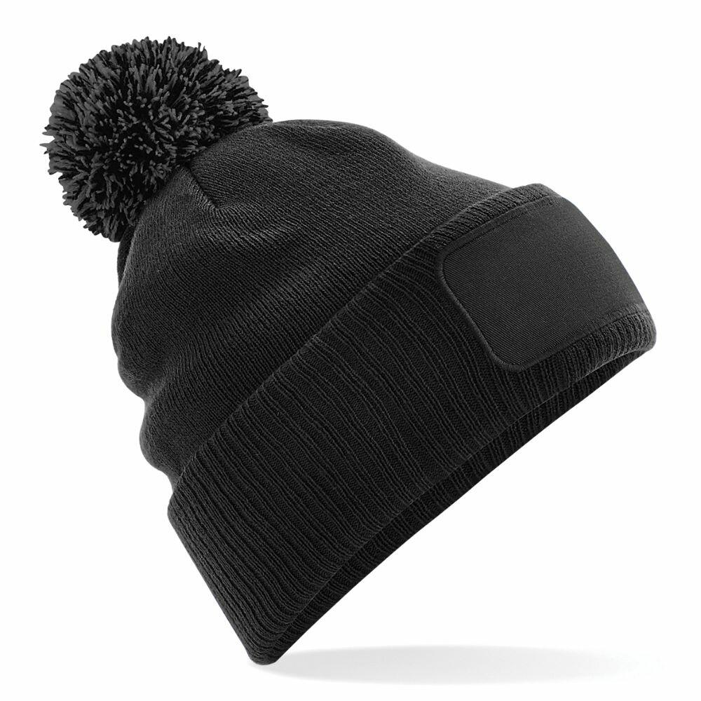 A Beechfield BC443 black knit beanie featuring a pom-pom on top and a blank rectangular patch on the front.