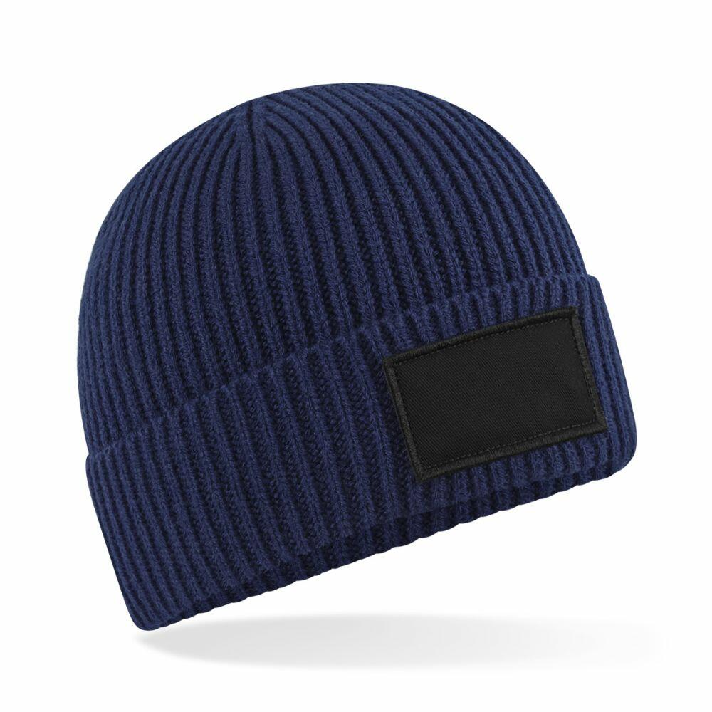 A Beechfield B442R blue ribbed knit beanie featuring a black rectangular patch on the front, positioned against a plain white background.