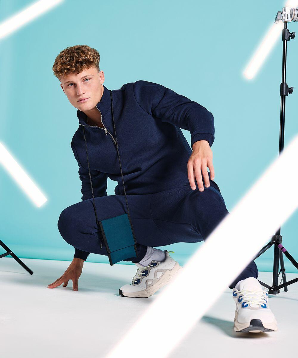 A young man with curly blonde hair crouches on one knee against a light blue background. He is dressed in a navy blue tracksuit and white sneakers. He wears a Bagbase BG283, and white light streaks are visible in the foreground. Two studio lights are also visible.