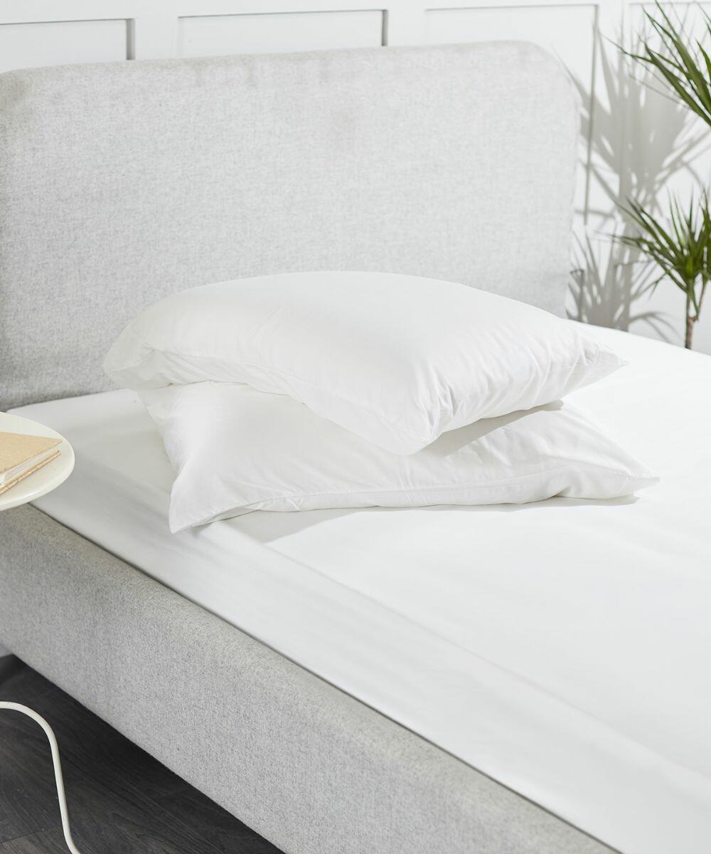 A neatly made bed with a white HL002 headboard by Home & Living, white sheets, and two white pillows stacked on top. A small table with a book and a plant is placed beside the bed.