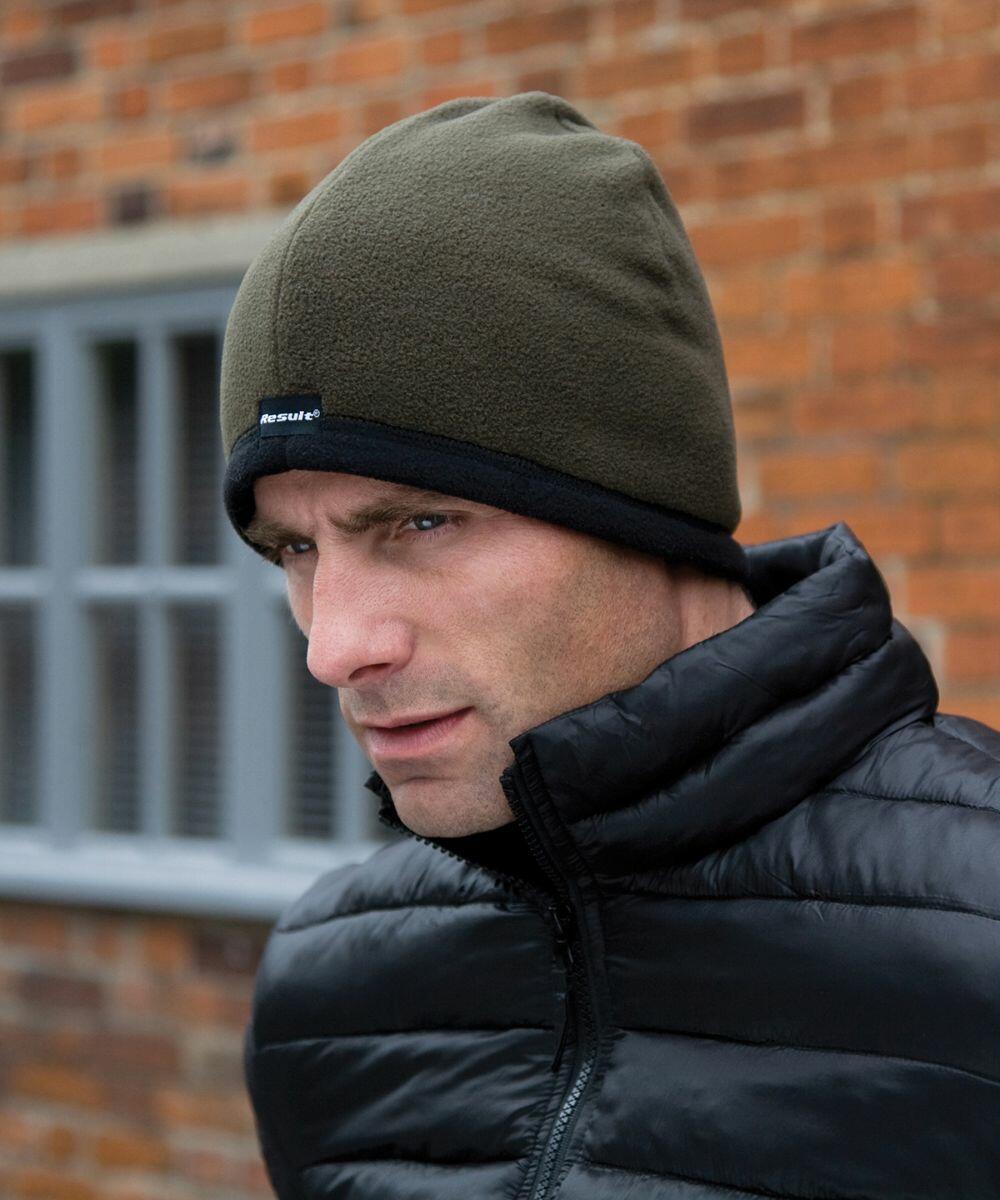 A man wearing a green beanie and a black Result RC142 puffer jacket stands in front of a brick wall with a window.