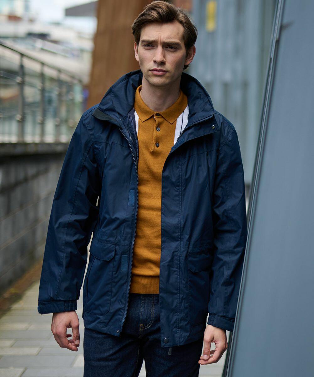 A person stands outdoors wearing a navy blue RG603 jacket by Regatta and a mustard yellow shirt, with an urban background featuring a metal railing and building.