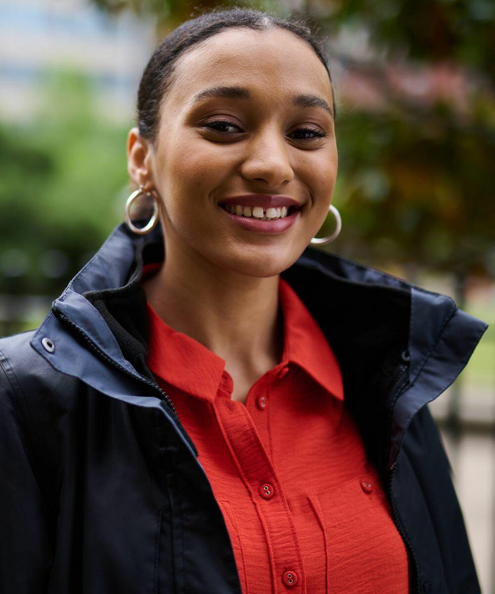 A woman in a red shirt and dark jacket stands outdoors, smiling. She has hoop earrings and her hair is pulled back. Trees and a building are blurred in the background, while she proudly wears the Regatta RG111.