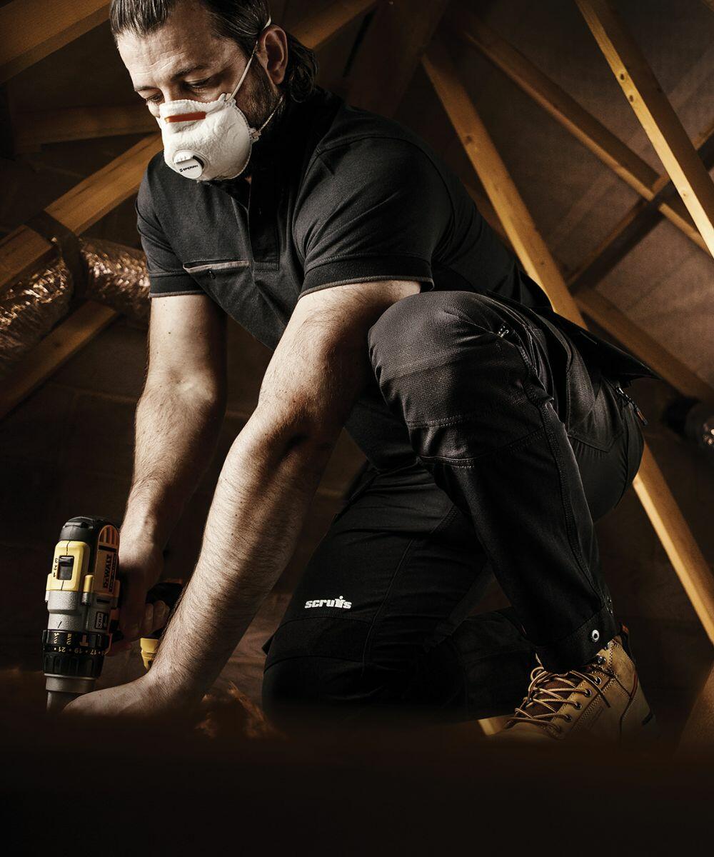 A worker in black clothing and safety mask kneels while using a Scruffs SH054 power drill in a construction site with exposed wooden beams.