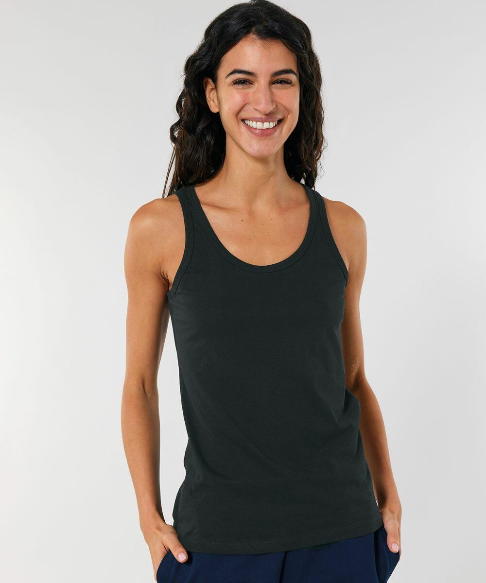 A person with long dark hair smiles, wearing a black SX013 tank top by Stanley/Stella and blue pants.