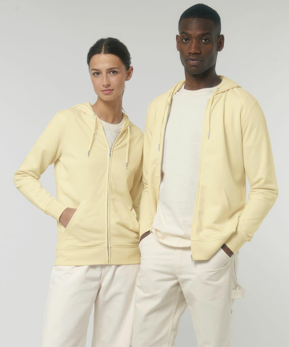 A woman and a man stand side by side wearing matching light yellow Stanley/Stella SX025 hoodies and white pants against a neutral background. Both have their hands in their pockets.