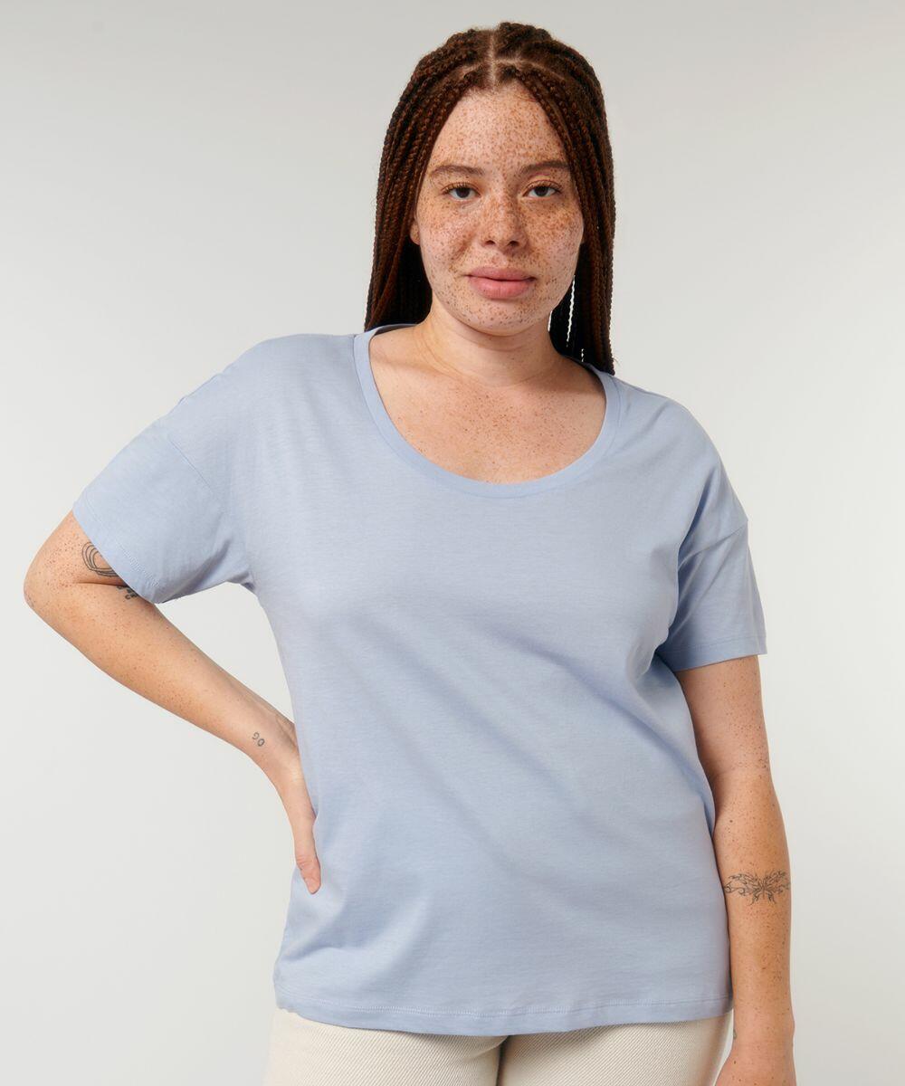 A person with long braided hair and freckles is wearing a light blue short-sleeve SX081 shirt from Stanley/Stella, standing against a plain light background.