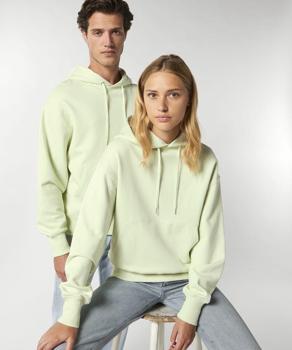 Two individuals wearing light yellow SX107 hoodies from Stanley/Stella; one is standing behind while the other is sitting on a stool. Both have neutral expressions and are facing forward.