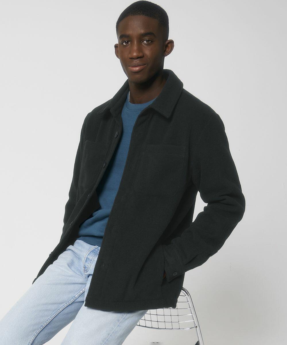 A man wearing a dark jacket over a blue shirt and light jeans sits on a wire chair against a plain backdrop, showcasing the SX205 from Stanley/Stella.