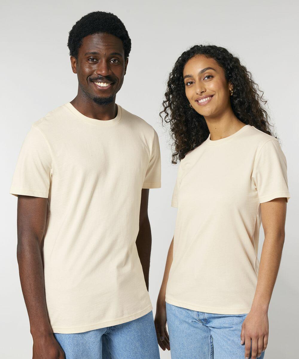 A man and a woman standing side by side, both smiling and wearing similar light beige SX236 T-shirts by Stanley/Stella and blue jeans, against a plain background.