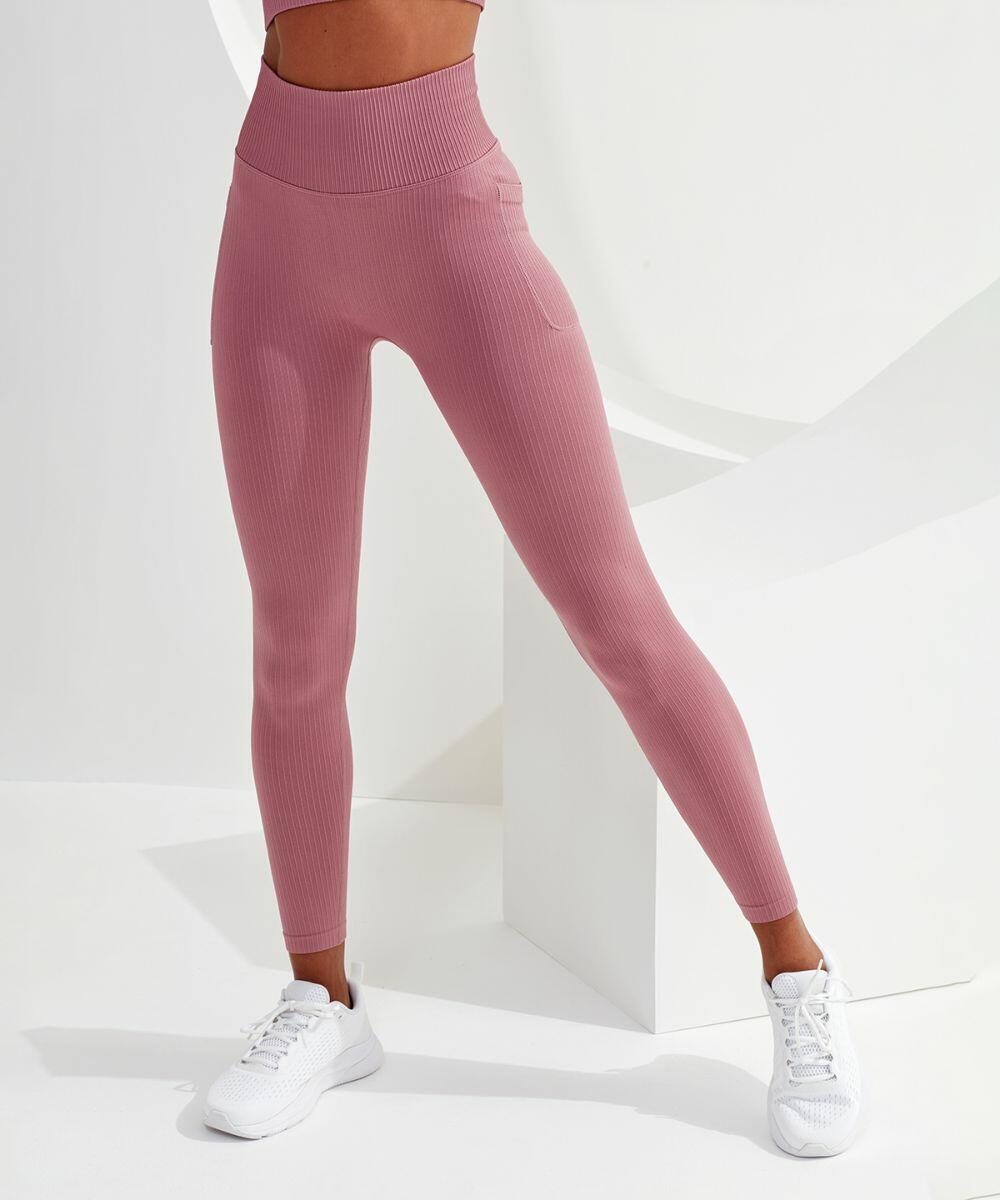 A person is wearing high-waisted, ribbed pink TR211 leggings by Tri-Dri and white athletic shoes. They are standing in a minimalist white space with a geometric background.