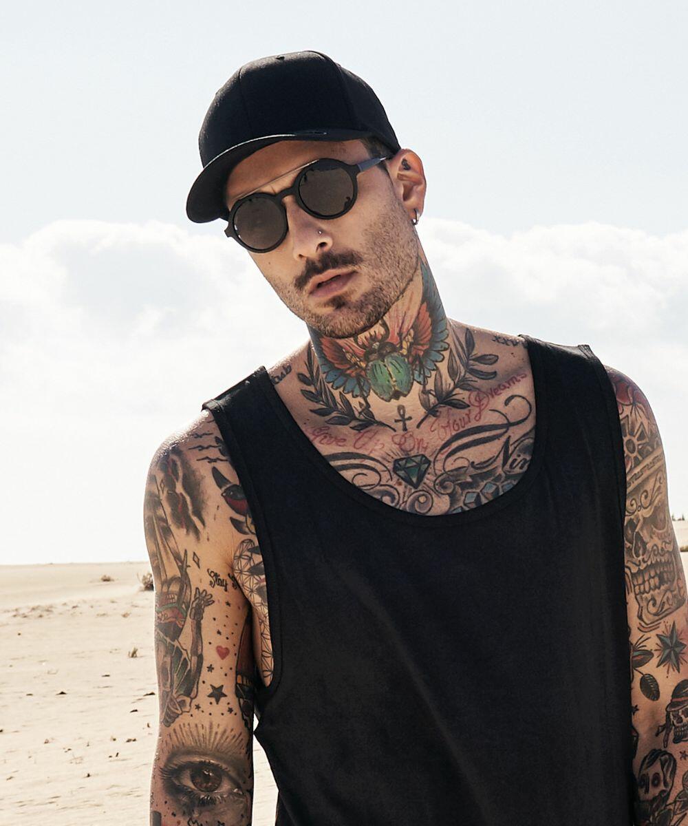 Man with tattoos wearing sunglasses, a black tank top, and a Flexfit By Yupoong YP222, standing in a desert-like area with a cloudy sky in the background.