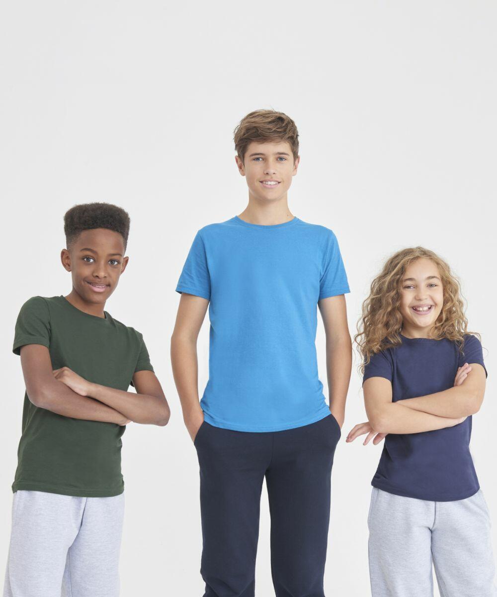 Three children stand side by side, each smiling and dressed in casual clothing. The child on the left wears an AWDis Ecologie EA01J shirt in green, the middle child wears an AWDis Ecologie EA01J shirt in blue, and the child on the right wears an AWDis Ecologie EA01J shirt in navy.