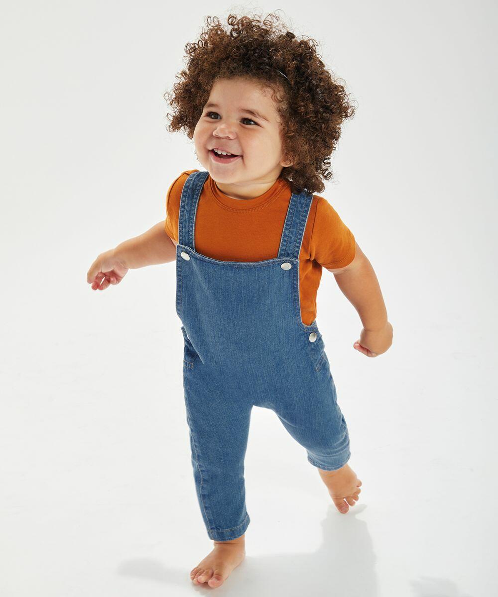 A smiling toddler with curly hair wearing a yellow BZ056 shirt and blue denim overalls from Babybugz walks barefoot on a white background.