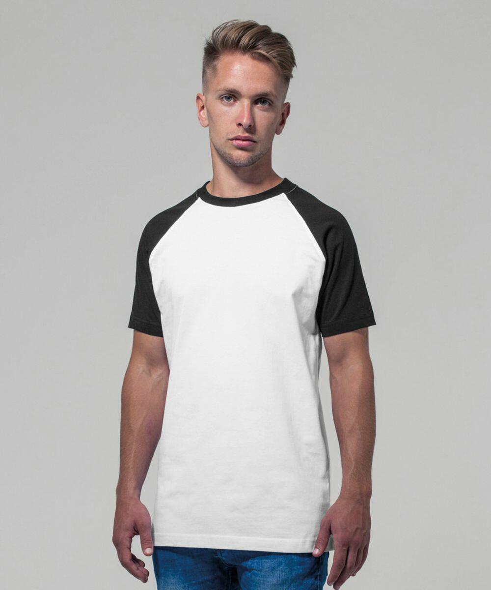 A person with short, light brown hair is wearing a white BY007 raglan t-shirt with black sleeves from Build Your Brand and blue jeans. They are standing against a plain, light gray background and looking directly at the camera with a neutral expression.