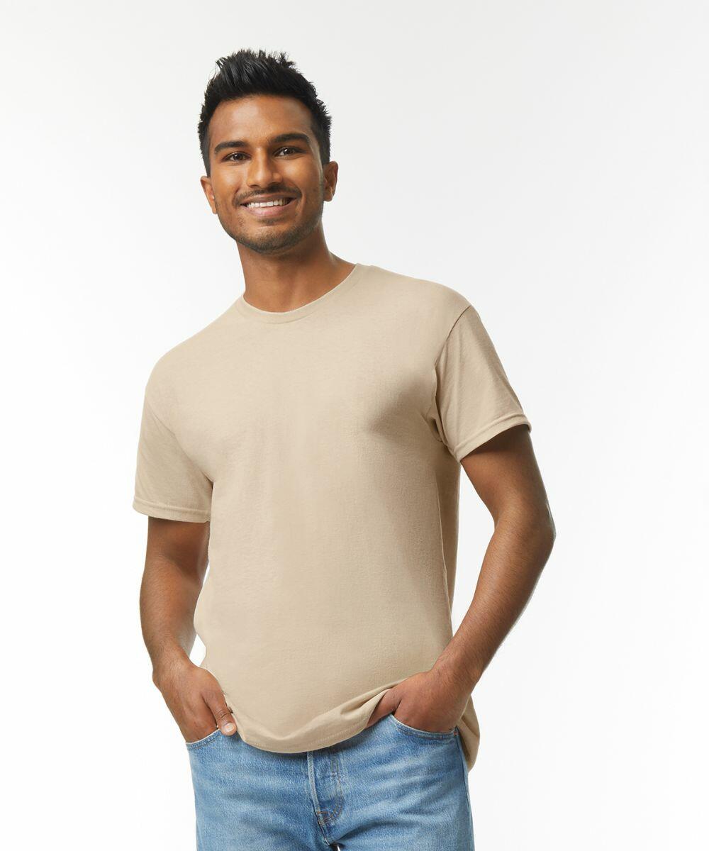 A person with short dark hair smiles while standing with hands in pockets, wearing a beige GD005 t-shirt by Gildan and blue jeans against a plain white background.
