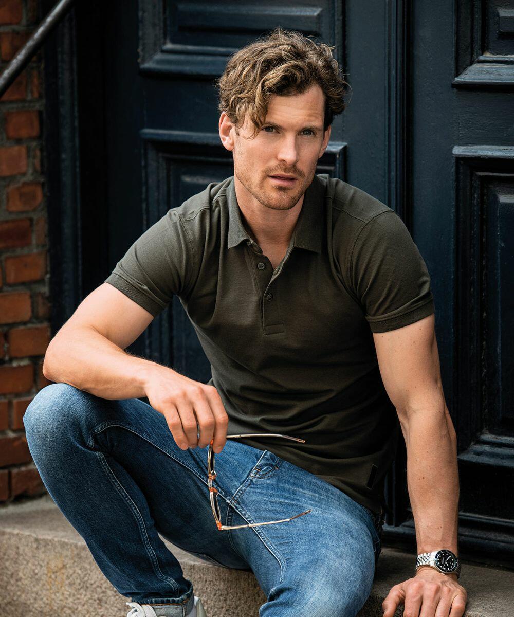 A man is sitting on a step, against a dark blue door, holding Nimbus NB52M eyeglasses in his hand. He wears a dark green polo shirt and blue jeans.