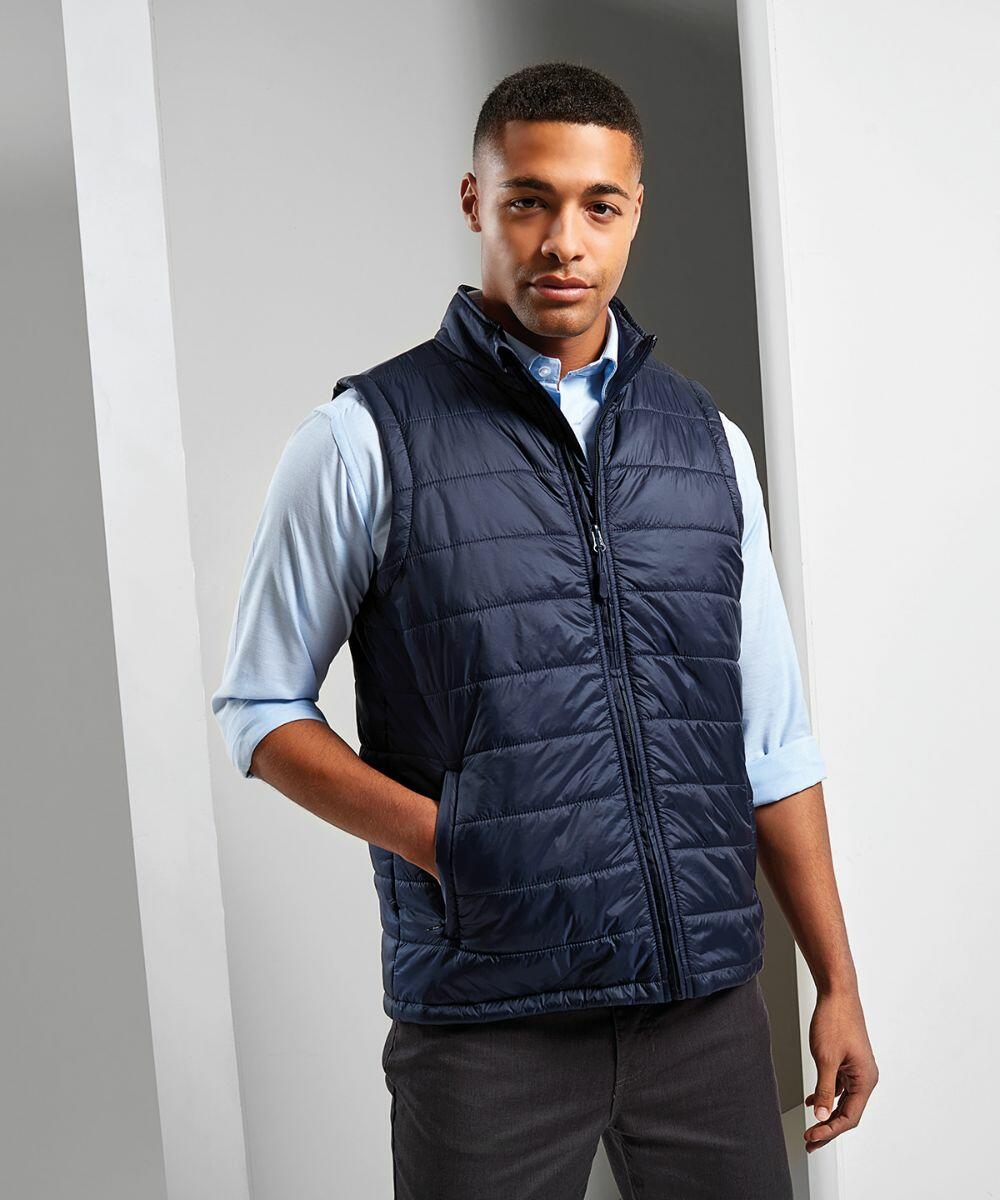 A man in a light blue shirt and a navy blue quilted Premier PR811 vest stands indoors with hands in his pockets.