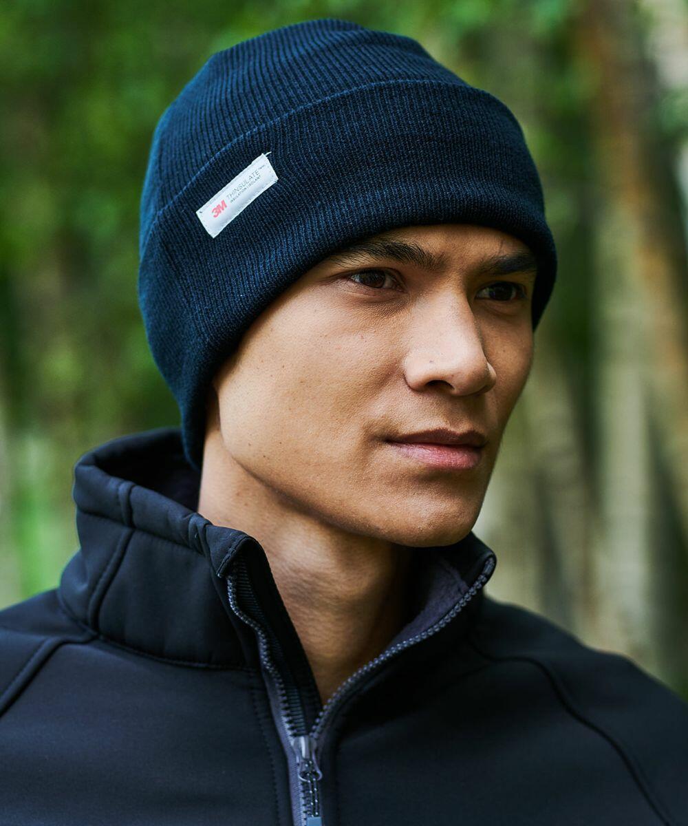 A person wearing a dark blue RG294 beanie by Regatta and a black zip-up jacket stands outdoors in front of a blurry green background.