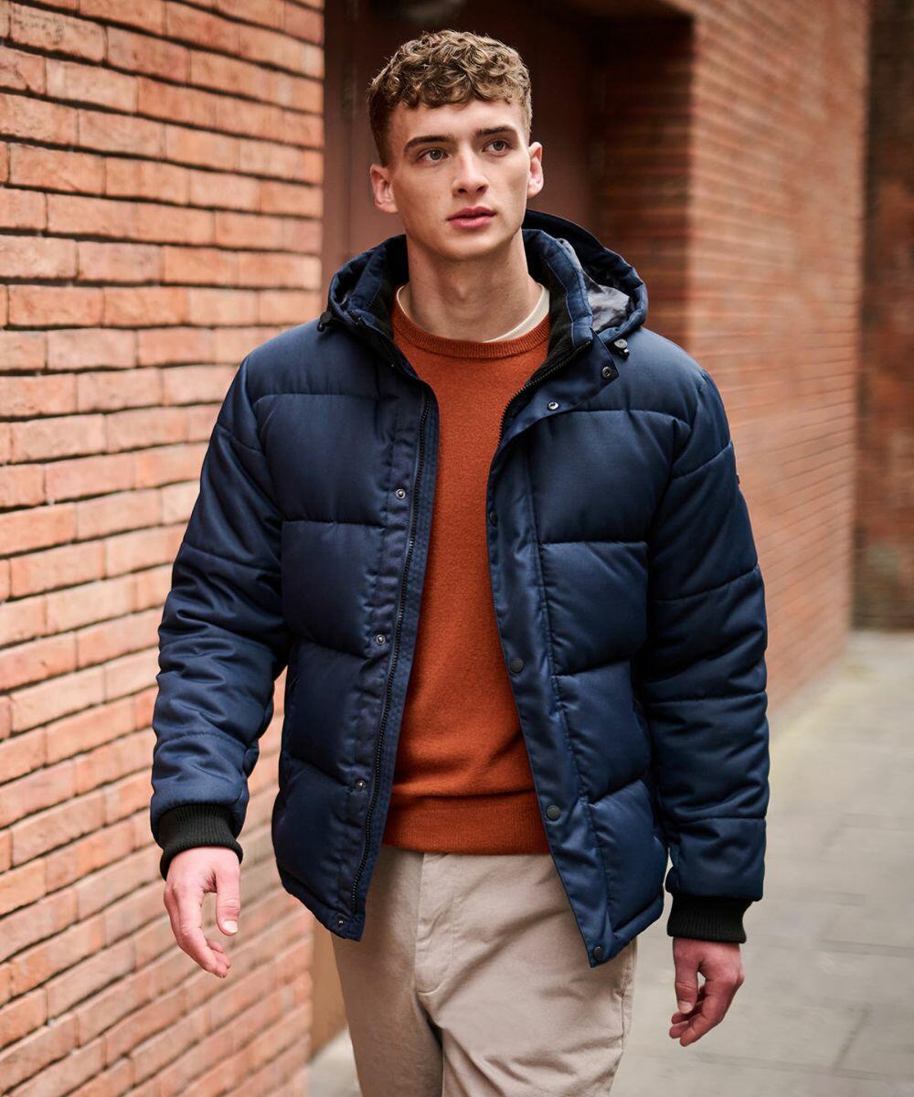 A man wearing a blue Regatta RG587 jacket, orange sweater, and beige pants walks beside a red brick wall.