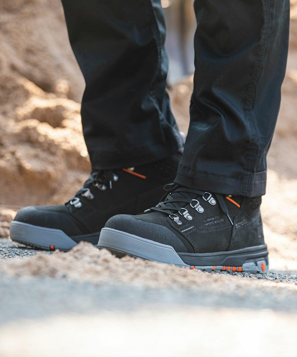 Man wearing black Scruffs SH081 work boots with orange accents standing on gravel near a sandy area.