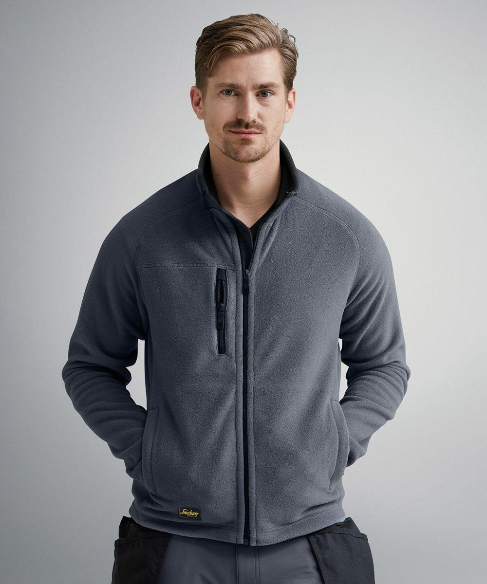 A man stands against a plain background wearing a gray zip-up fleece jacket from Snickers and black SI036 pants.
