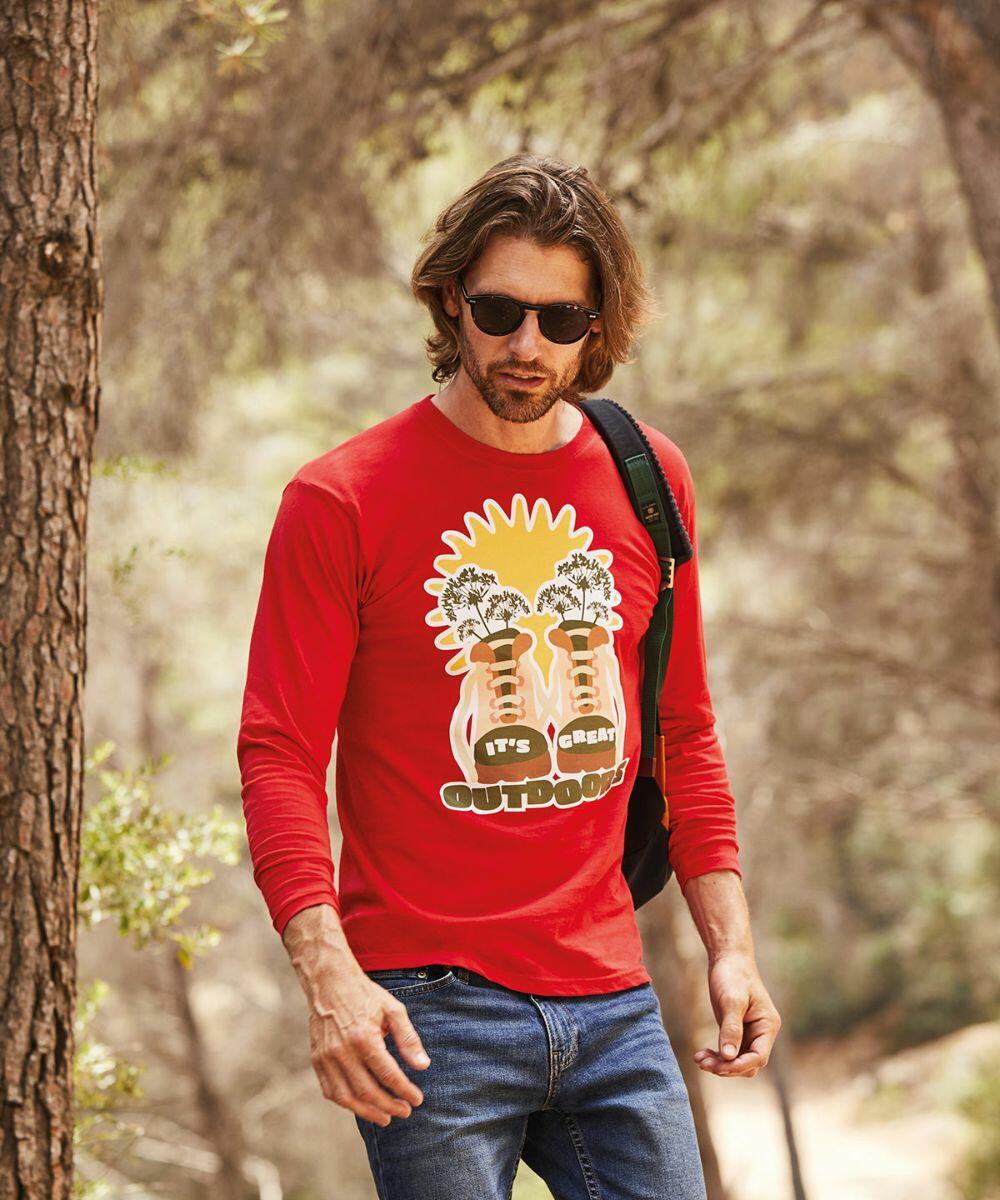 A man with long hair wearing sunglasses, a Fruit of the Loom SS032 red outdoor-themed graphic T-shirt, and blue jeans is walking in a forest with a backpack.