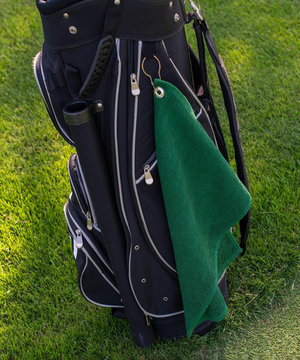 A black golf bag with multiple zippered compartments stands on grass. A green TC013 from Towel City is hanging from a hook on the side of the bag.