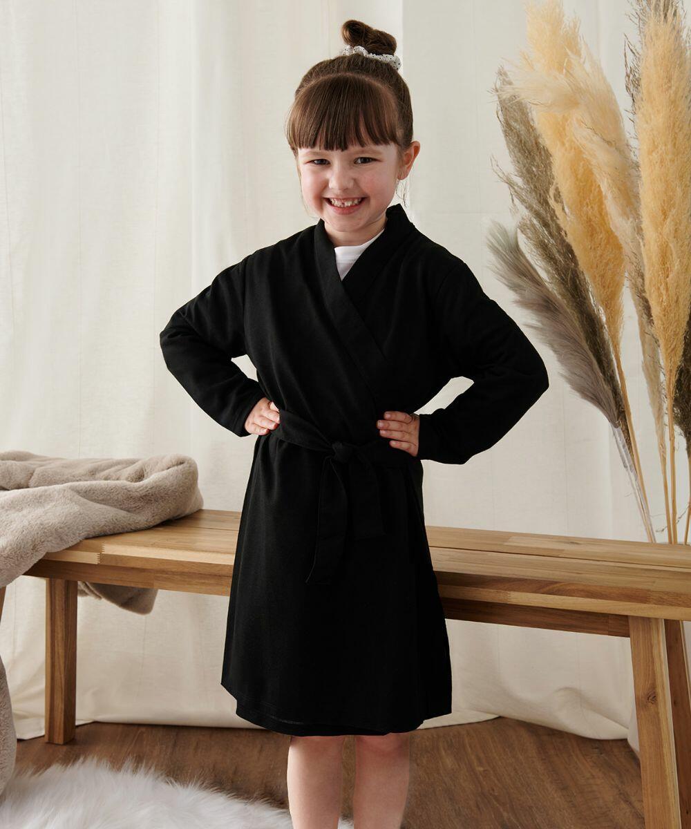 A young girl with a fringe and a bun smiles while standing with her hands on her hips. She is wearing TC051 by Towel City, a black dress with a tied waist. A wooden bench and decorative plants are in the background.