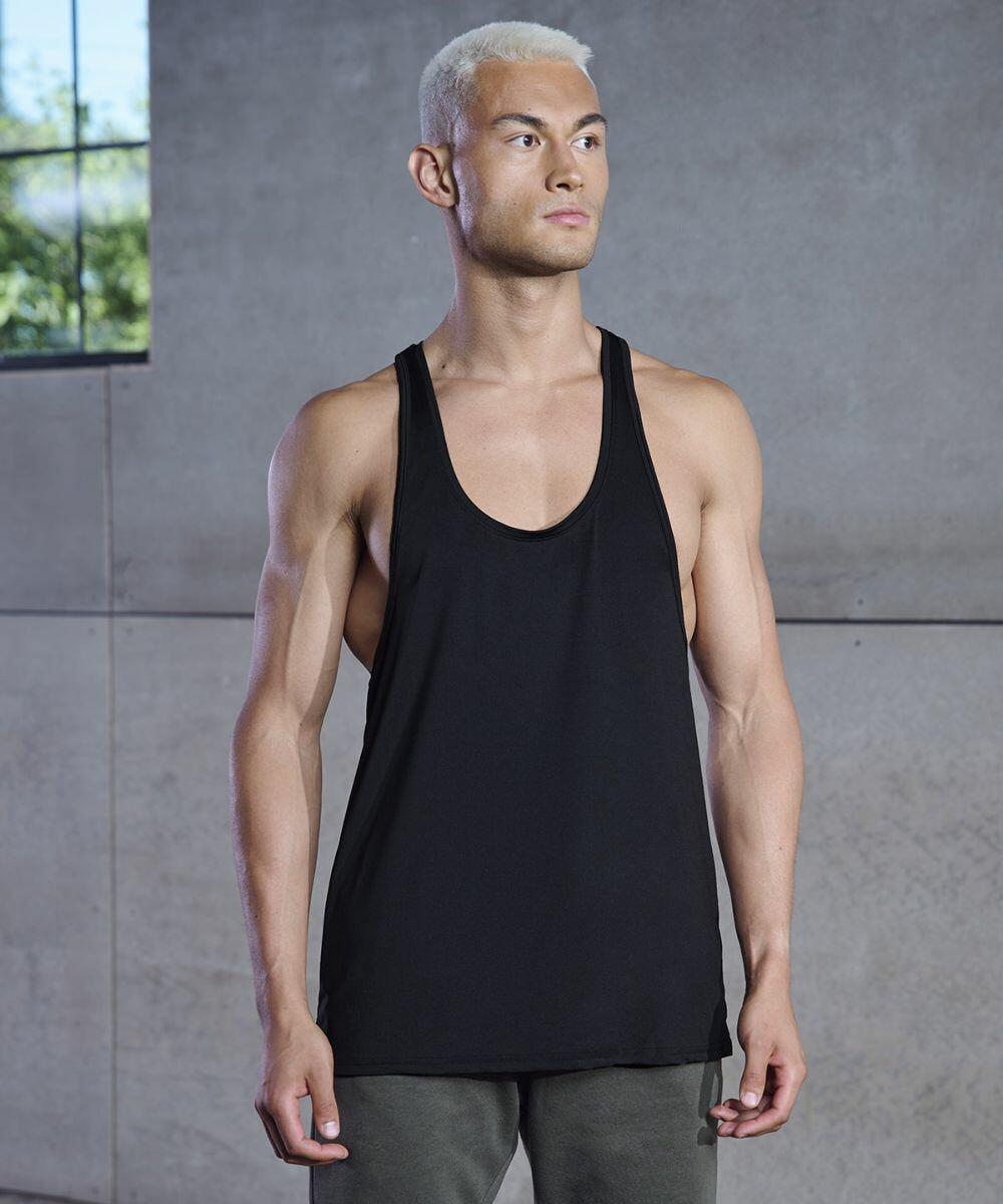 A person with short blonde hair stands in front of a window in a concrete room, wearing a Tombo TL504 black tank top and dark pants.