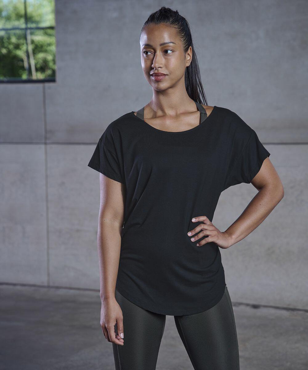 A woman in Tombo TL527 wears stands indoors by a concrete wall, looking to the side with a relaxed expression. She has her hair tied back and one hand on her hip.