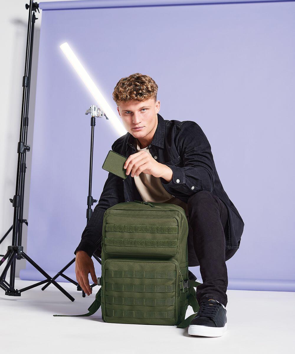 A young man with short curly hair is posed in a studio with a purple backdrop, holding a green phone case. He's wearing a black jacket, pants, and sneakers. He rests one hand on a Bagbase BG840 placed in front of him and studio lighting equipment is visible in the background.