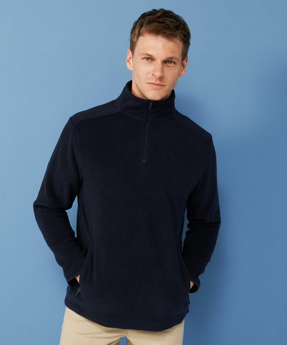 A man stands against a blue background wearing a dark Henbury HB858 pullover fleece jacket with a half-zip front and beige pants, hands in pockets.