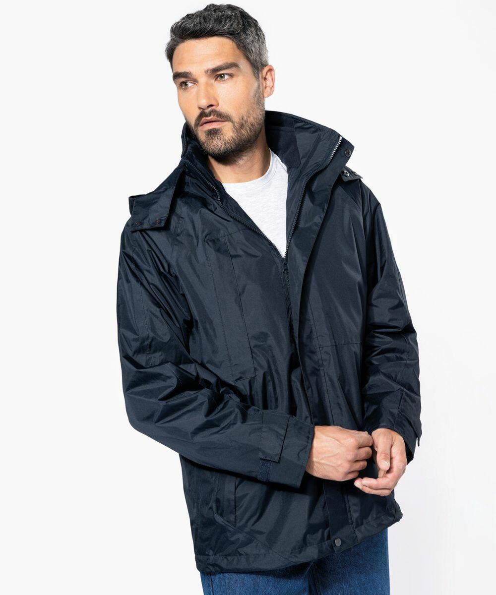 A man with short hair and a beard is wearing a dark blue Kariban KB657 rain jacket, white shirt, and blue jeans. He is looking slightly to his left, with one hand adjusting the zipper of his jacket.