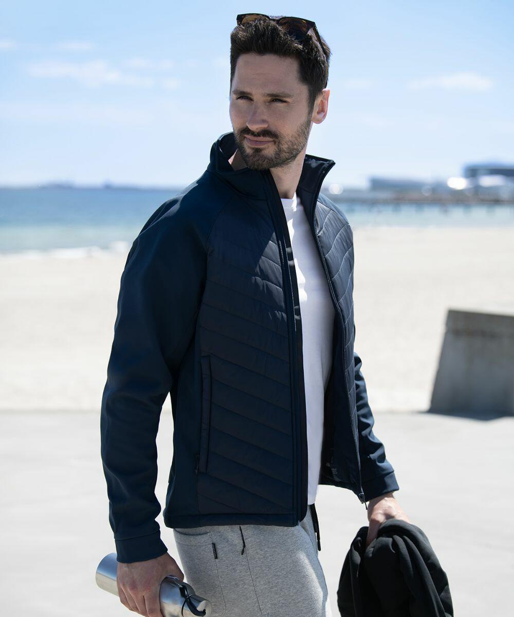 A man in a blue jacket and gray pants stands on a beach holding an NP09M by Nimbus and a piece of clothing. The background features sand, a pier, and the ocean.