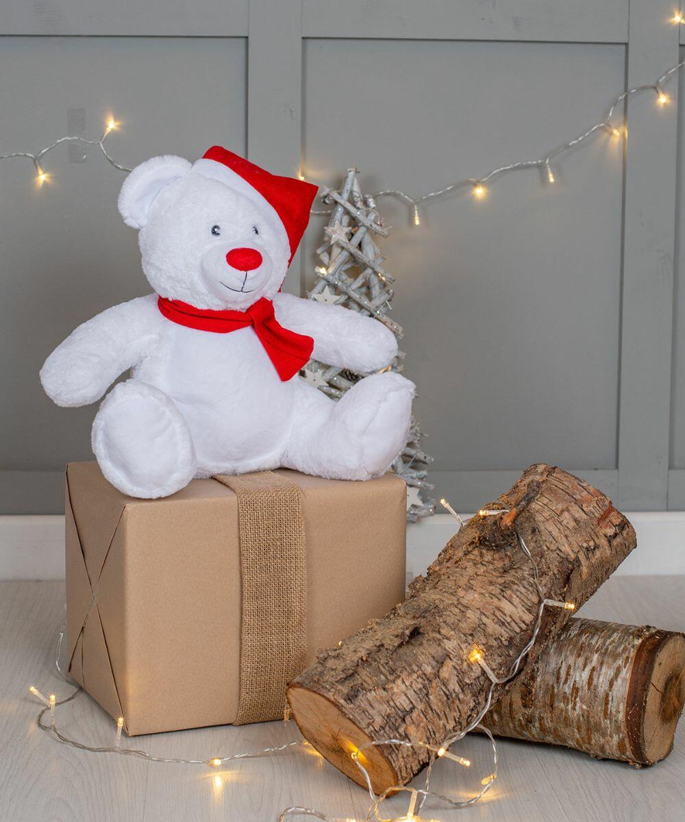 Mumbles MM573 with a red Santa hat and scarf sits on a wrapped gift box next to two logs wrapped in fairy lights. Background includes a small white tree and decorative light strings.