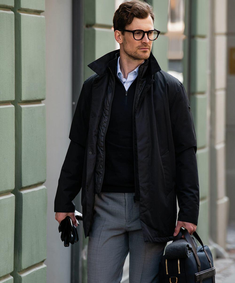 A man with glasses, wearing a black jacket over a black sweater, carries a Nimbus N105M and a pair of gloves while standing on a city street.