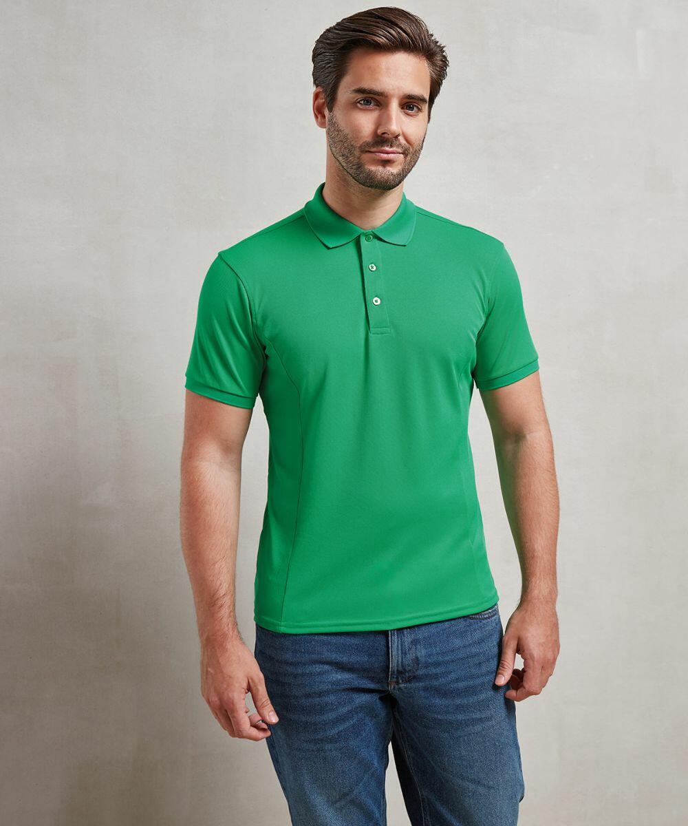 A person standing and wearing a green Premier PR630 short-sleeve polo shirt and jeans, posed against a plain, light-colored background.