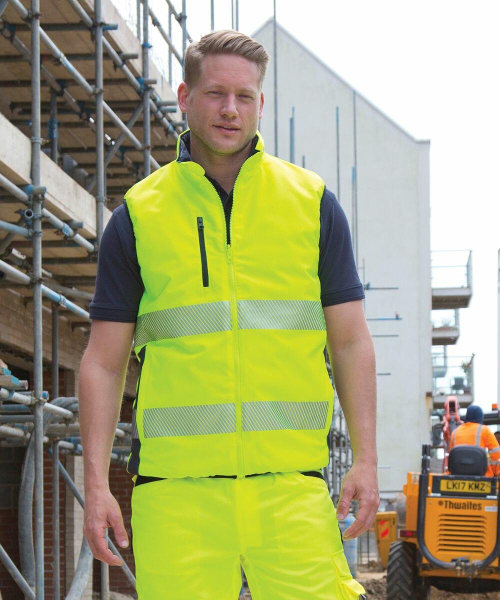 A man wearing a high-visibility yellow Result R332X safety vest and pants stands on a construction site near scaffolding and machinery.