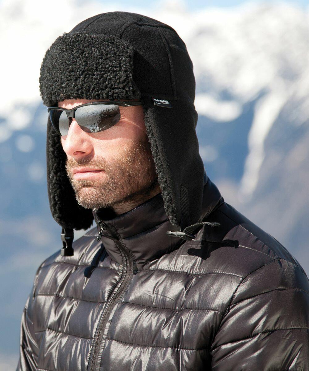 A person wearing sunglasses and a black Result R358X winter hat with ear flaps and a black puffer jacket, stands against a snowy mountain background.