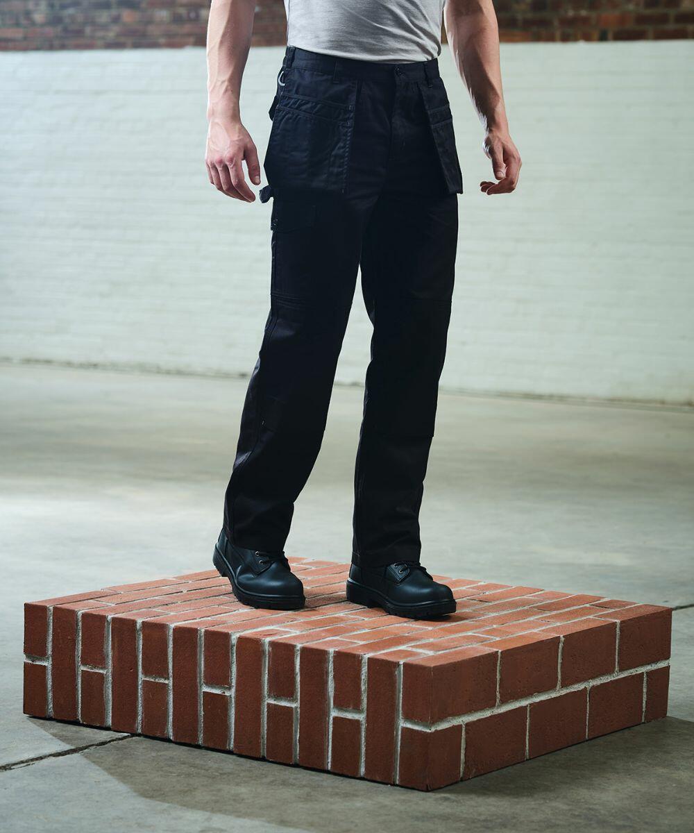 Person standing on a platform made of bricks wearing black pants and black boots, with a plain white and brick-colored wall background, showcasing the RG323 by Regatta.