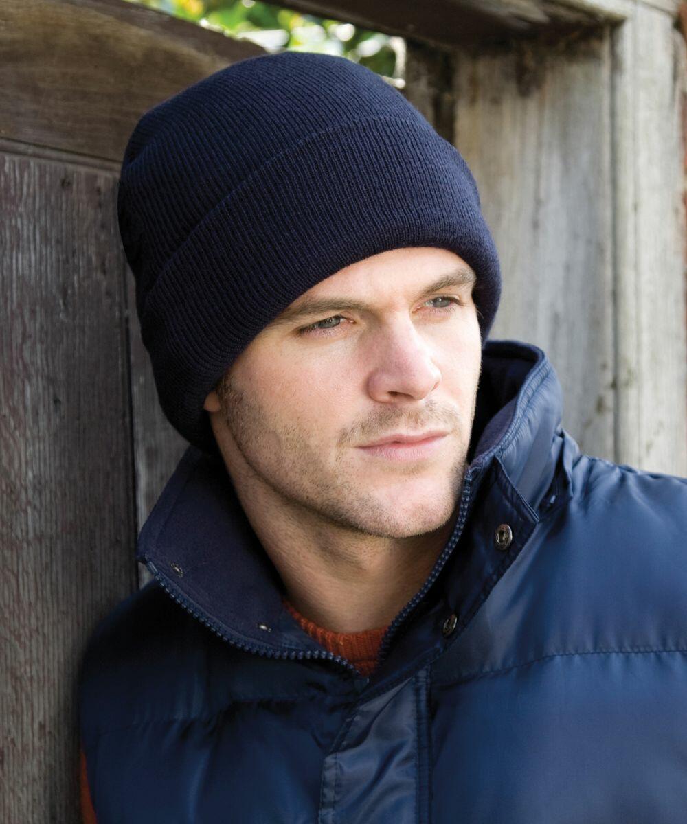 A man wearing a navy blue knit hat and a blue padded jacket from Result, model RC033, leans against a wooden wall, looking into the distance.
