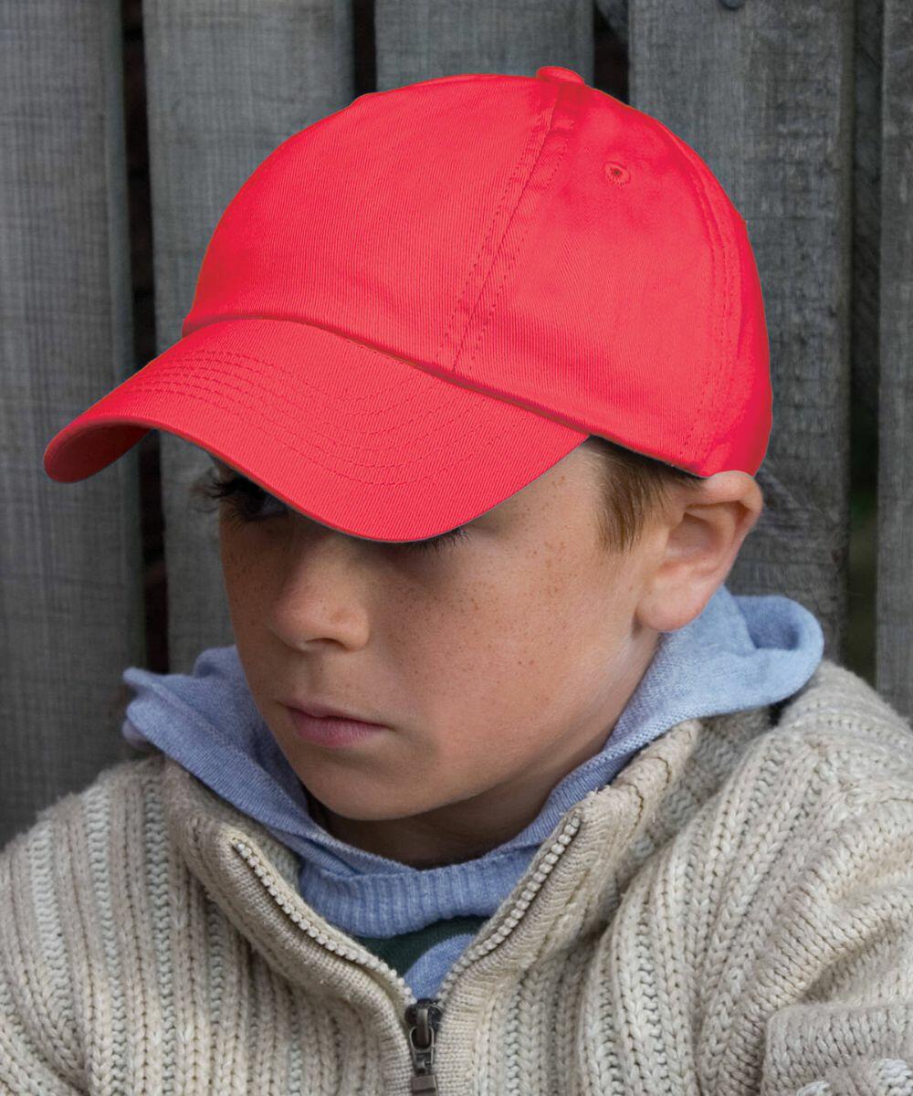 A young boy wearing a red cap, a grey hoodie, and a beige knitted sweater sits with his arms crossed in front of a wooden fence, sporting the RC18J by Result.