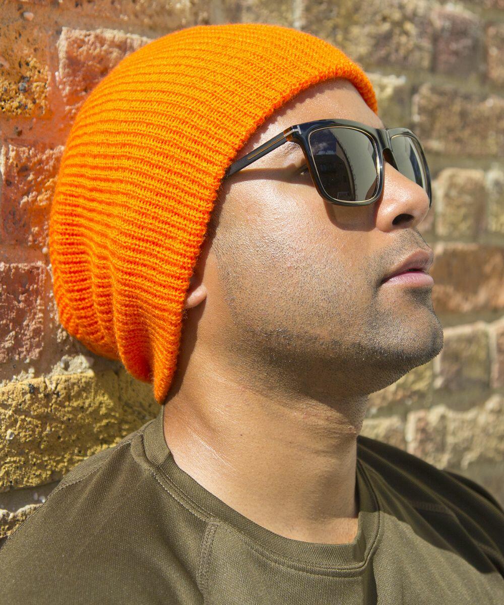 Person wearing an orange Result RC31X beanie and black sunglasses is leaning against a brick wall, looking up.