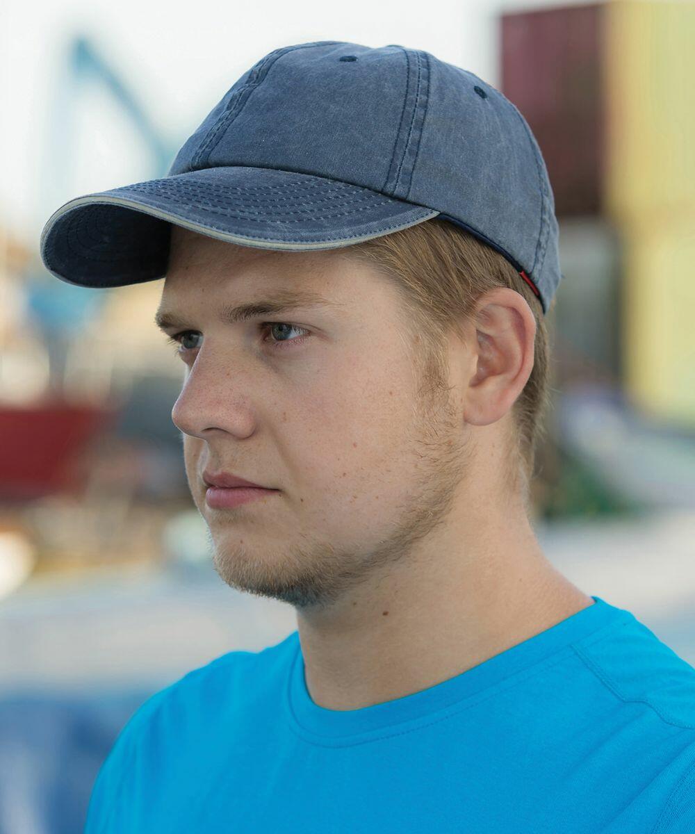 A young man wearing a blue cap and a Result RC54X blue shirt looks to the side. The background appears to be an outdoor setting with blurred structures.