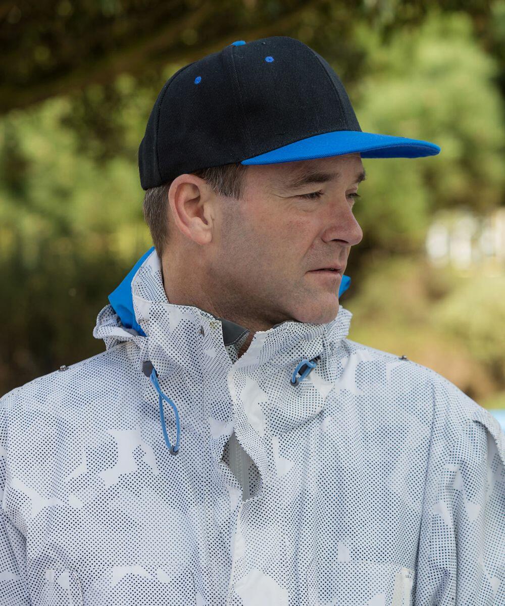 A man wearing a dark baseball cap and a light-colored, patterned RC82X Result jacket looks to his right with a neutral expression. Background is blurred greenery.
