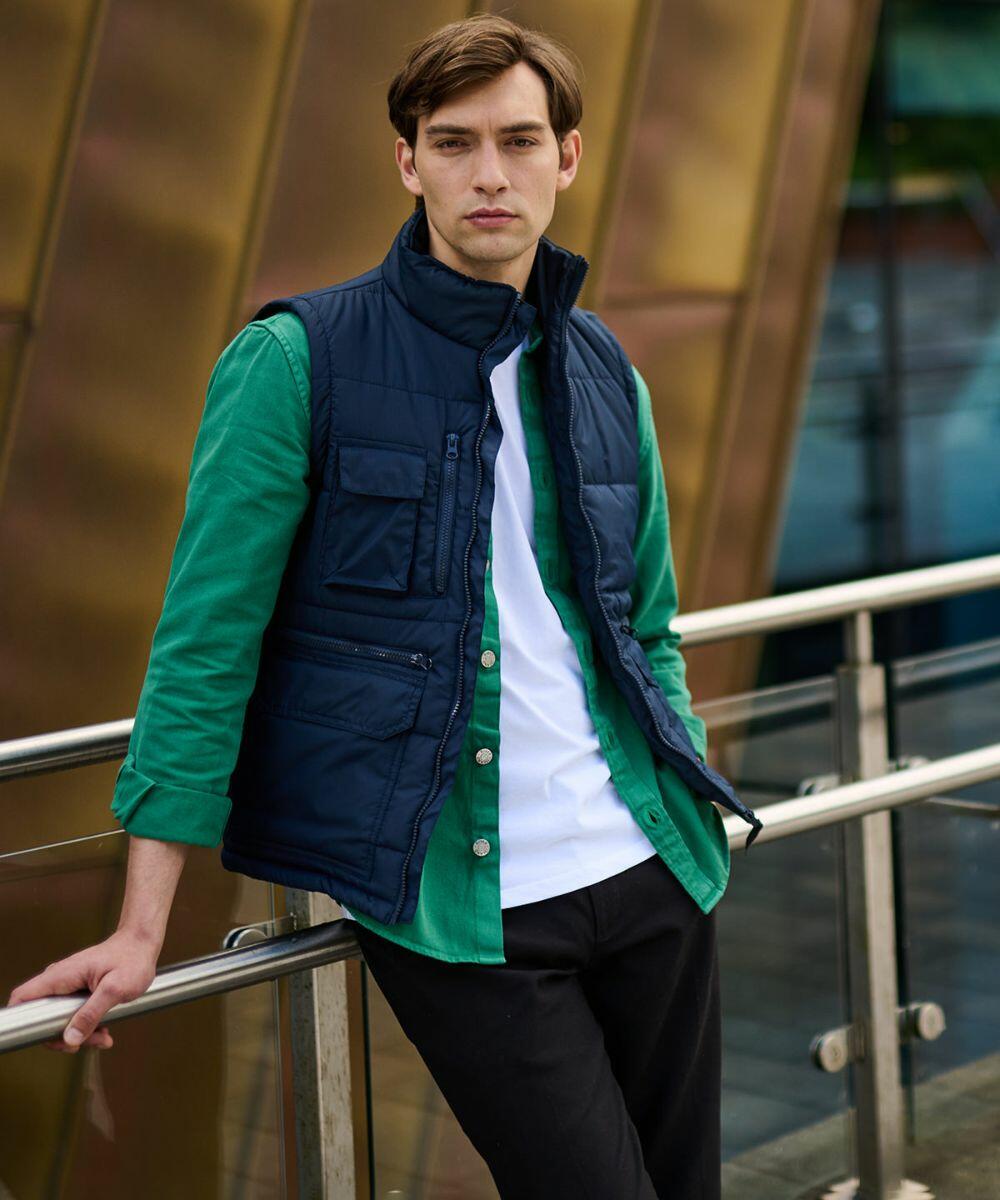 Man leaning against a rail, wearing a green shirt with a white tee underneath and an RG074 blue padded vest by Regatta, looking towards the camera. Background features an architectural structure.