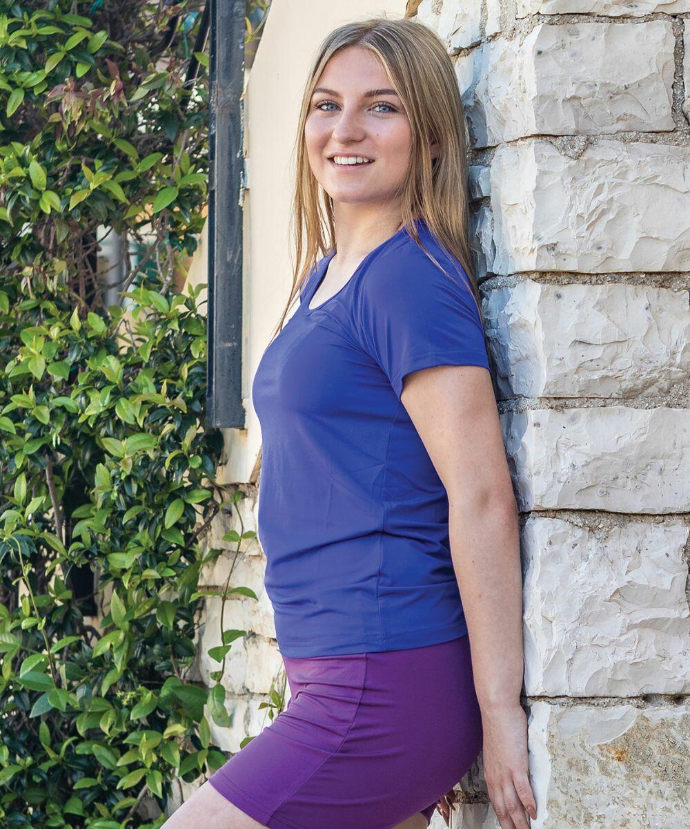 A person with long hair, wearing a blue Spiro S280F shirt and purple shorts, stands leaning against a stone wall. Green foliage is visible in the background.