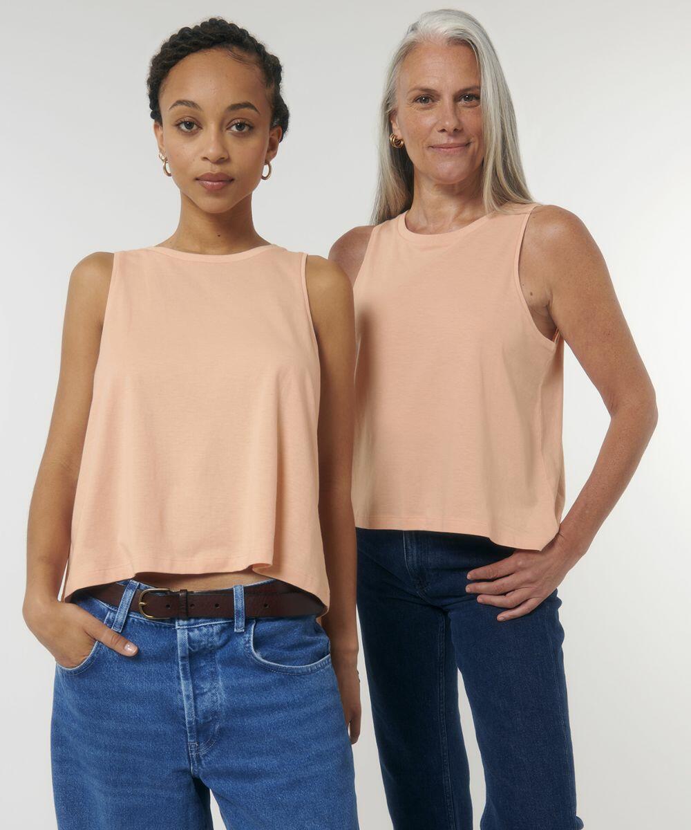 Two women, one younger and one older, stand side by side wearing Stanley/Stella SX082 peach sleeveless tops and blue jeans. Both look directly at the camera with a neutral expression, hands in pockets.
